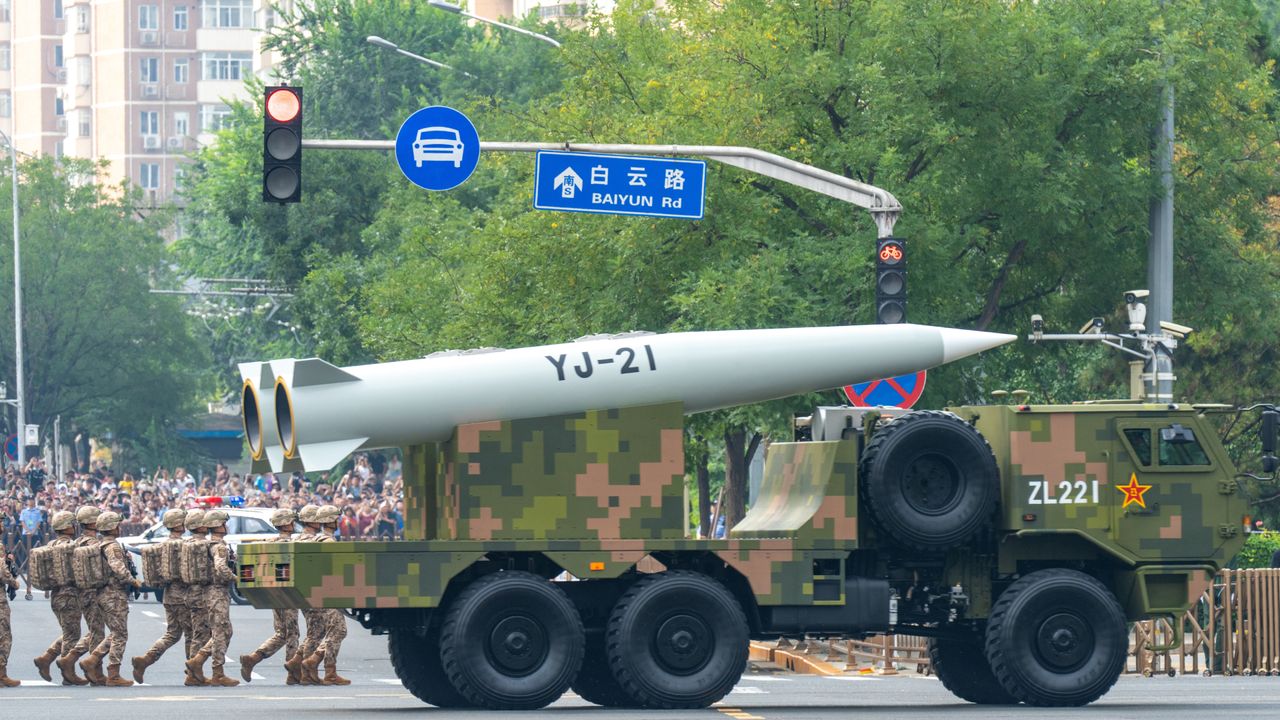 A large military truck carrying a huge hypersonic missile turns right at an intersection next to a series of marching officers