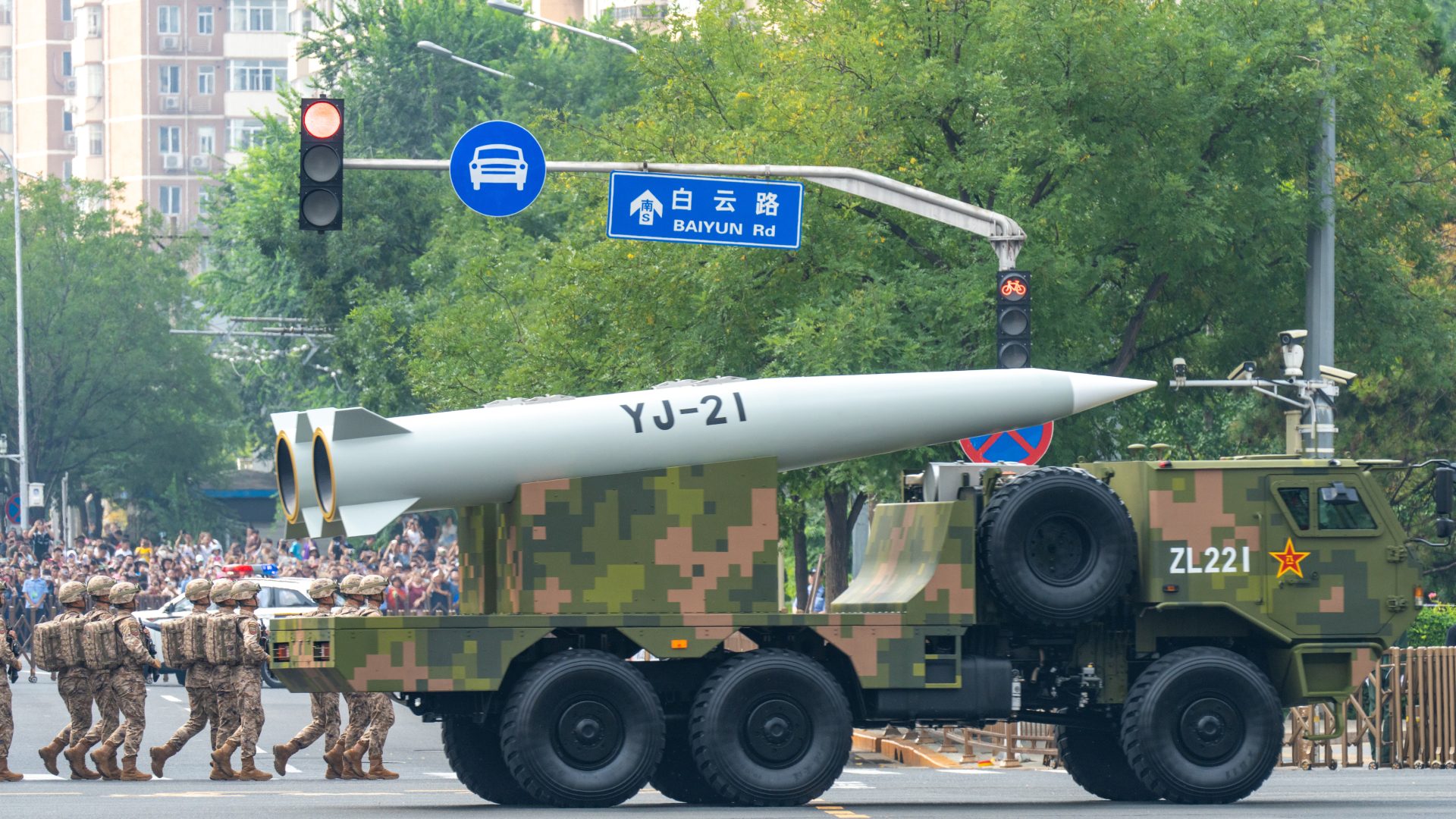A large military truck carrying a huge hypersonic missile turns right at an intersection next to a series of marching officers