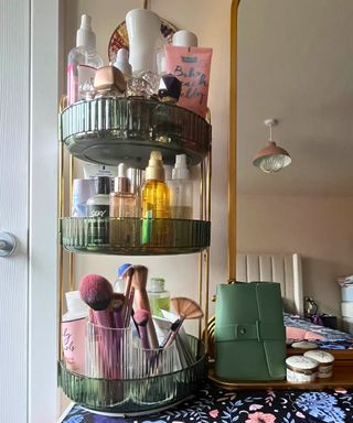 Gold ornate flower crown mirror on right reflecting tall cream headboard and to the left, a green fluted three tier organizer stacked with beauty and hair products plus makeup brushes