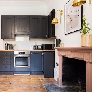 Kitchen with charcoal cupboards and golden handles next to fireplace with pink mantelpiece and a pot plant on top of it