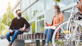 two college students sit outside on either end of a bench, both are wearing face masks and have laptops and books out