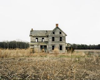 A weathered, gray wooden two-story farmhouse stands derelict and decaying in the middle of a barren, harvested cornfield under an overcast sky.