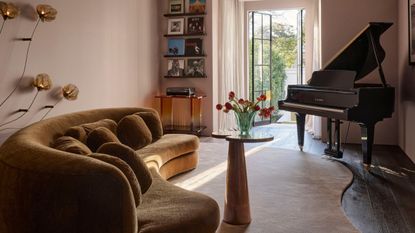 pink living room with curving rug, curved yellow sofa, wall light, tall side table with vase of flowers, piano, and shelves on wall with records displayed and a record player beneath, and french doors