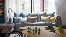 An airy living room space with large windows, sheer white curtains, pale blue couch with mismatched patterned cushions, a freestanding lamp, patterned upholstered stool, and a large coffee table in the foreground with green candle holders, three green bowls, and two green glasses.