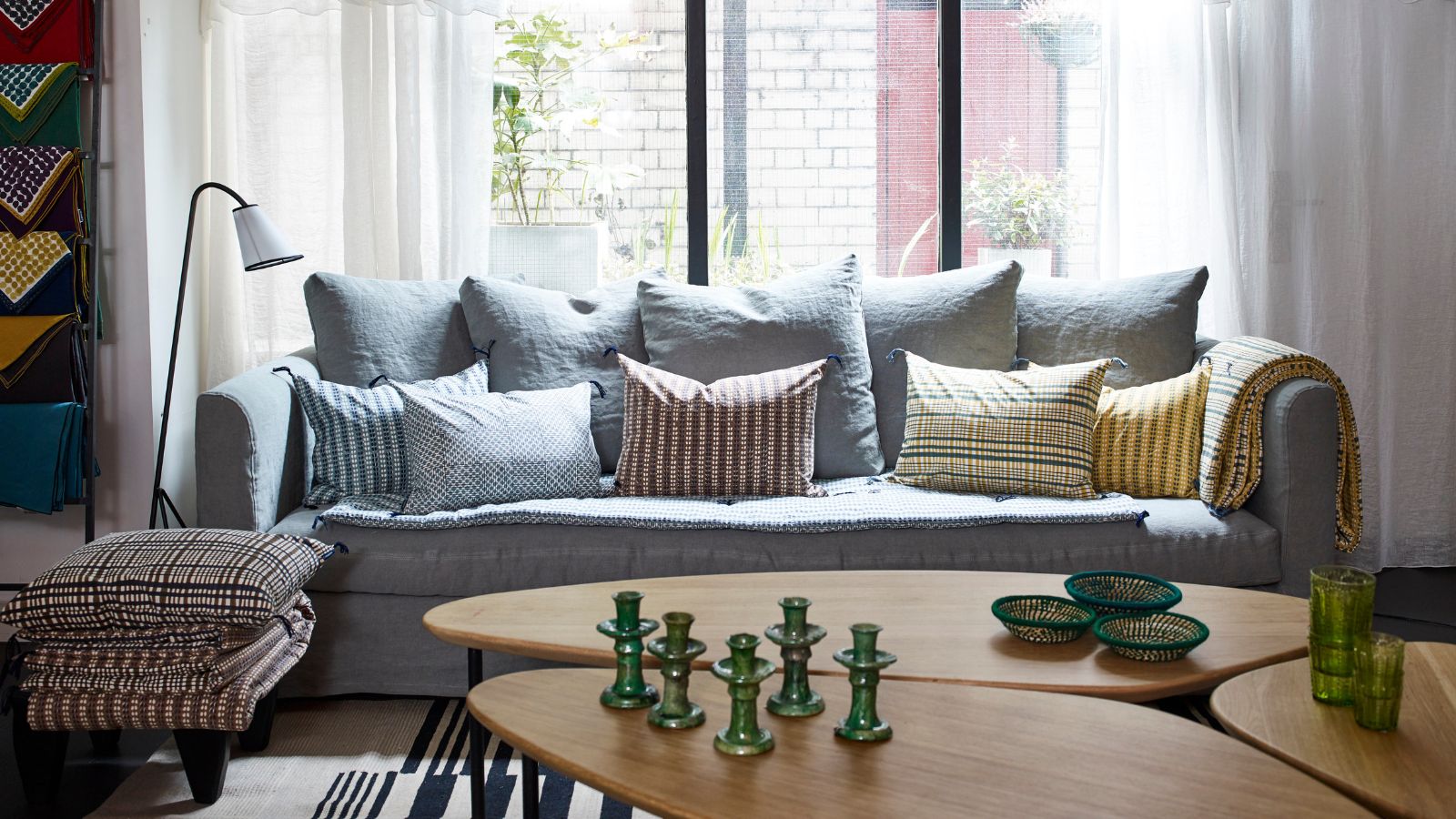 An airy living room space with large windows, sheer white curtains, pale blue couch with mismatched patterned cushions, a freestanding lamp, patterned upholstered stool, and a large coffee table in the foreground with green candle holders, three green bowls, and two green glasses.