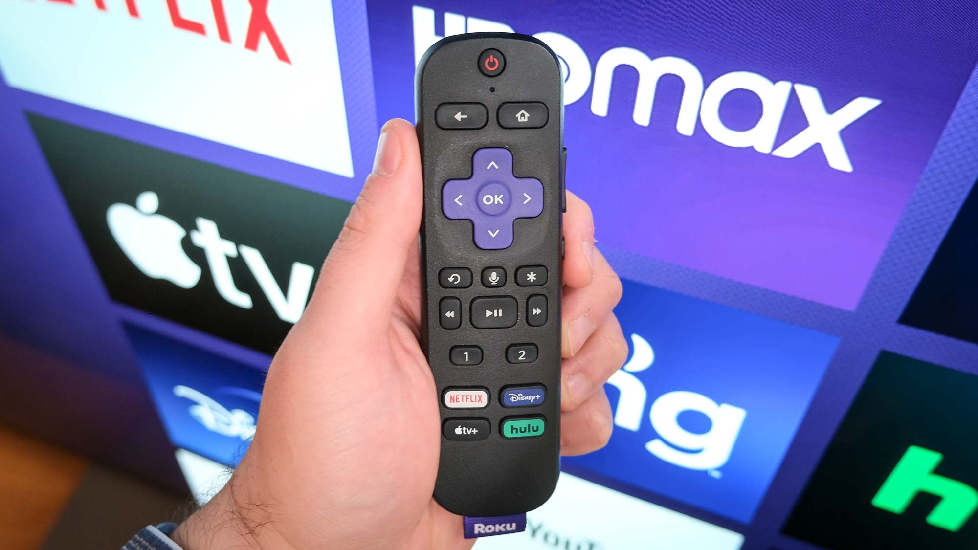 A Roku remote in hand in front of a TV with the Roku homepage.