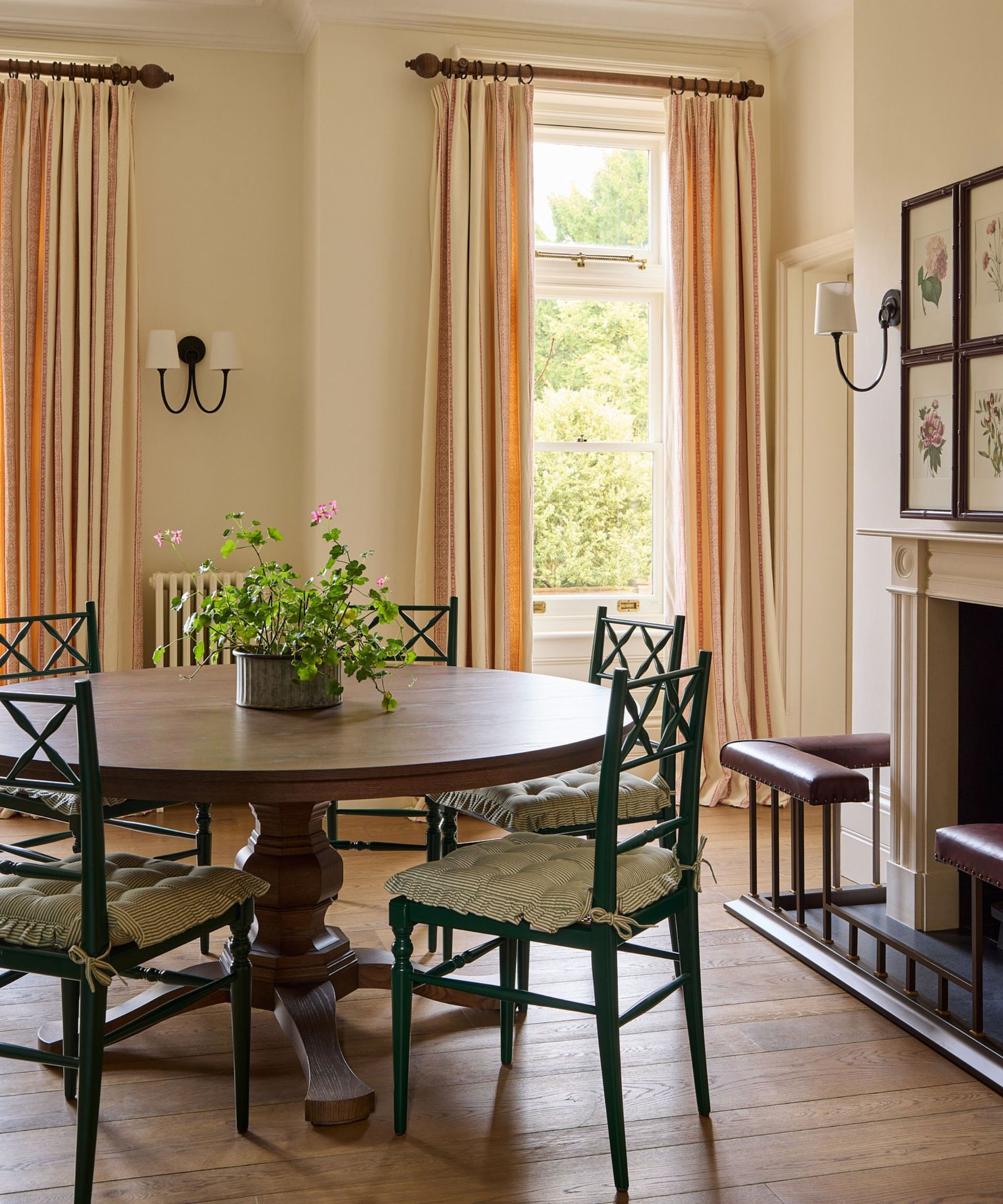 Round wooden dining table paired with dark green lattice-back chairs in a bright, cream-colored room with tall windows and striped curtains.