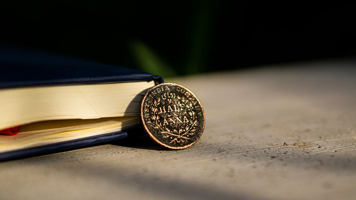 An ancient East India Company coin on a desk next to a leather-bound book