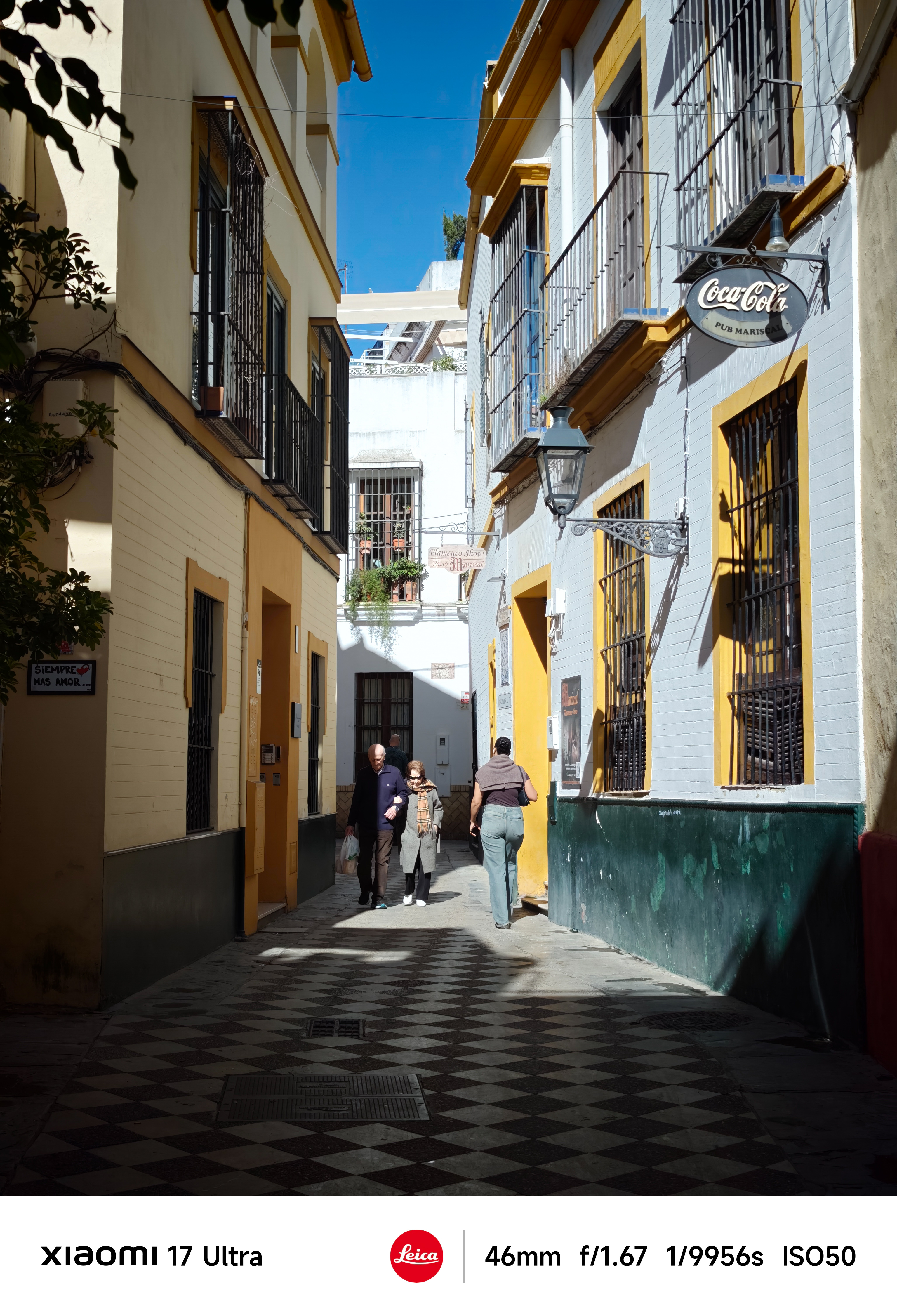 Narrow sunlit alley with pastel buildings and pedestrians walking along a patterned tiled floor.