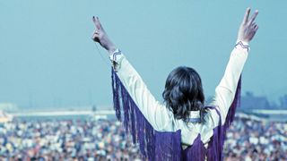 Ozzy onstage at the California Jam in 1974, back to the camera, arms raised in triumph, saluting the audience