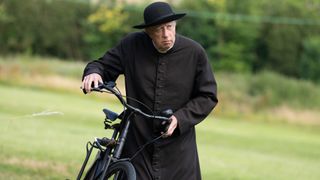 Father Brown (Mark Williams) in a black robe walking besides a bicycle