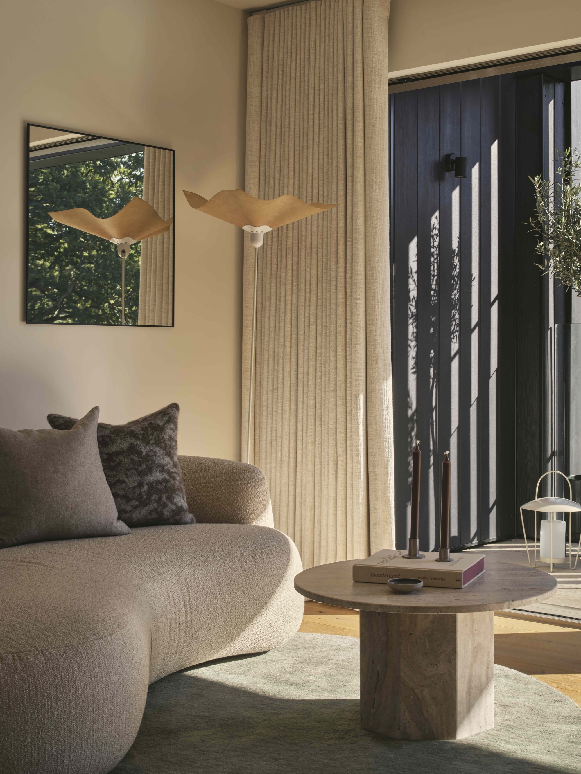beige living room with curtains, round green rug, with travertine coffee table, gray curved sofa with cushions, and raffia pendant light and square mirror