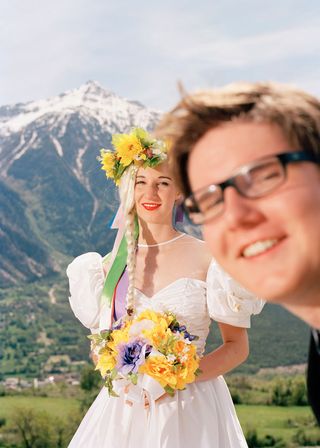 Young blonde woman in a white wedding dress, flower tiara, pearl necklace, holding a bouquet of yellow, blue and white flowers, man in glasses at the frongt of the shot, snowy mountainous landscape and village in the backdrop, cloudy pale blue sky