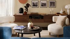 Two curved white boucle armchairs sit either side of a round dark wood coffee table in a living room with a blue sofa just out of view and a wall hung console with pictures above and decor on top