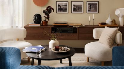 Two curved white boucle armchairs sit either side of a round dark wood coffee table in a living room with a blue sofa just out of view and a wall hung console with pictures above and decor on top