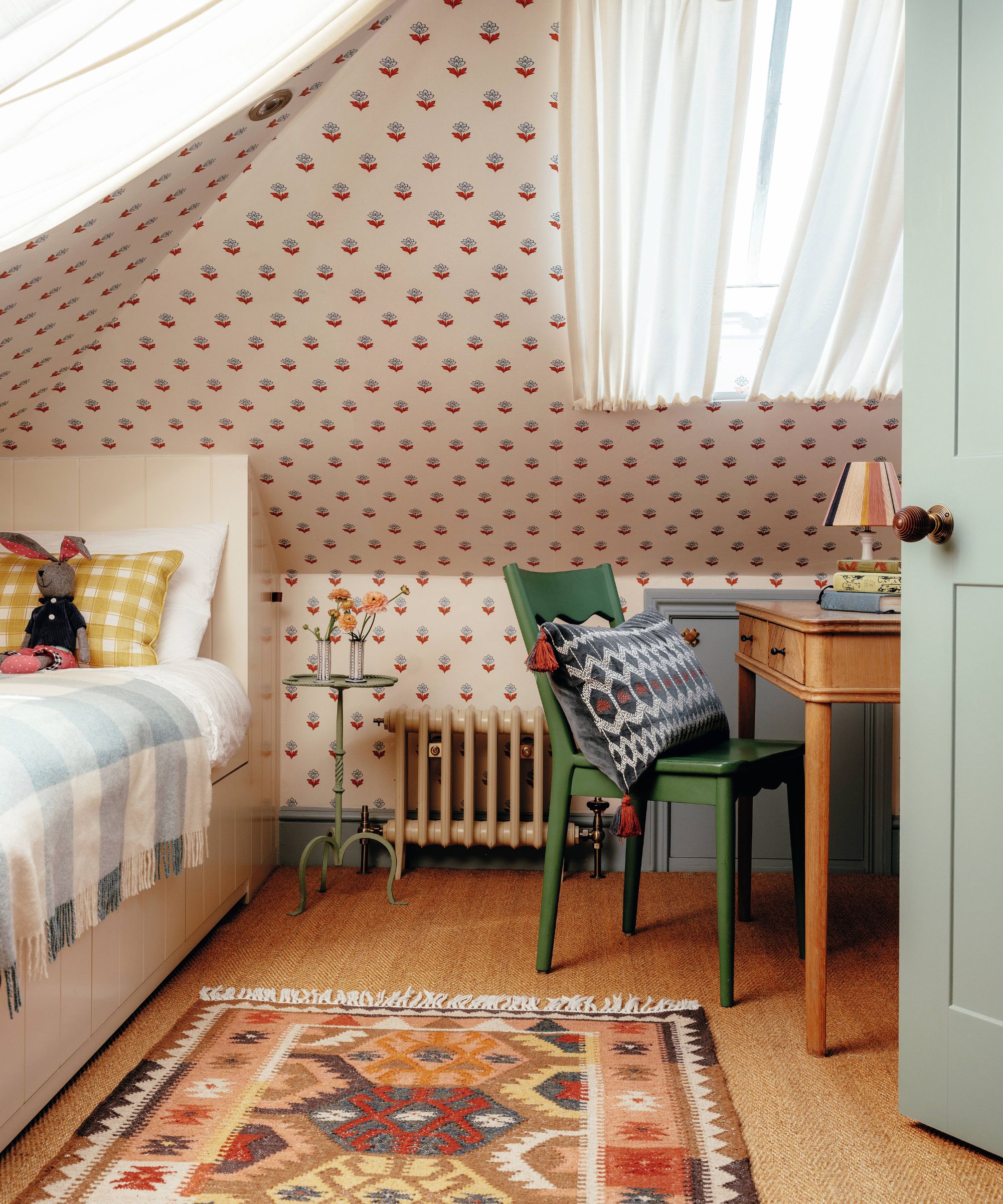 an attic kids bedroom with floral wallpapered walls and ceiling, green painted trim, a built in single bed and a small wooden desk with green chair