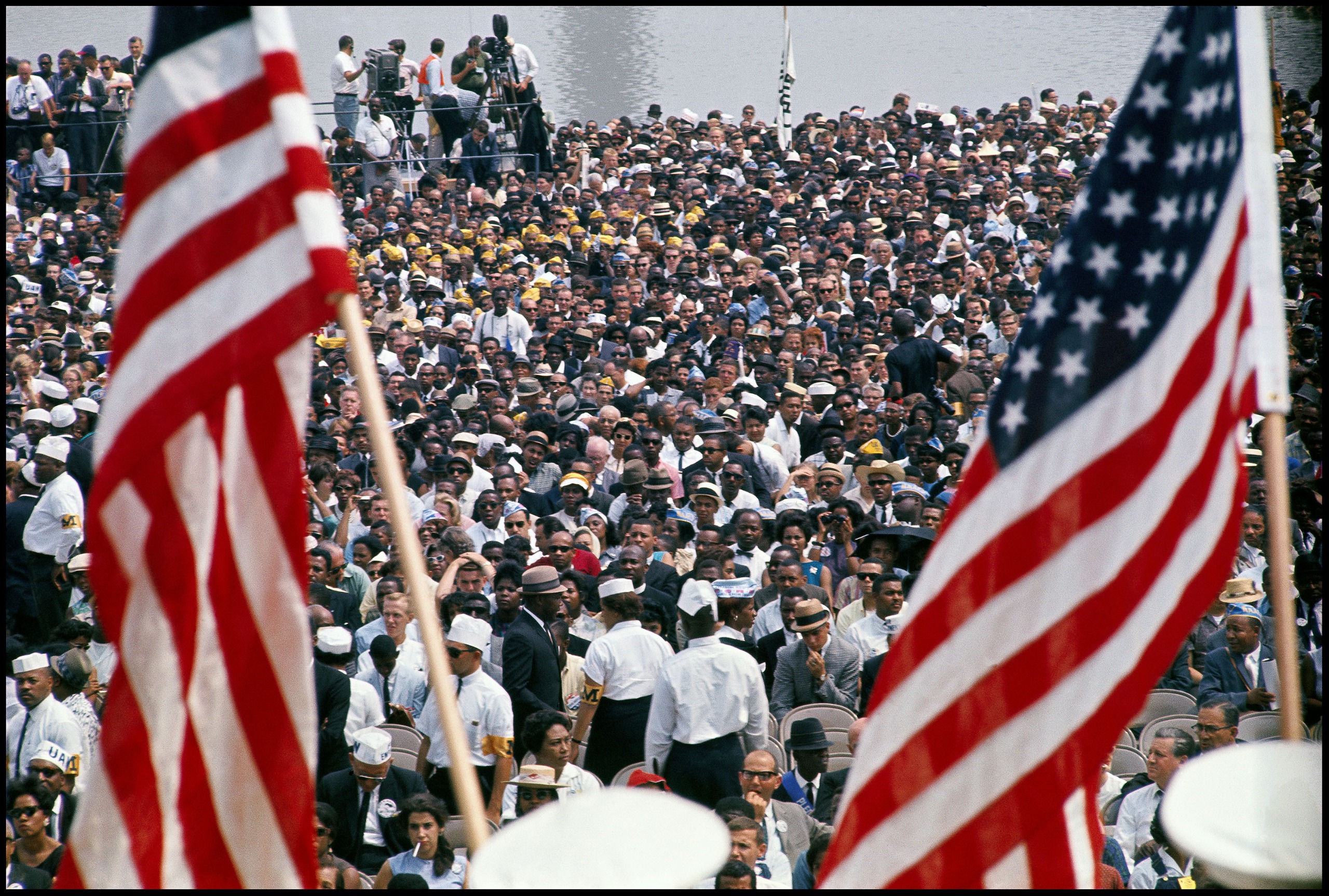 A dense crowd fills the frame at the 1963 March on Washington, with two American flags held in the foreground against a backdrop of thousands of demonstrators