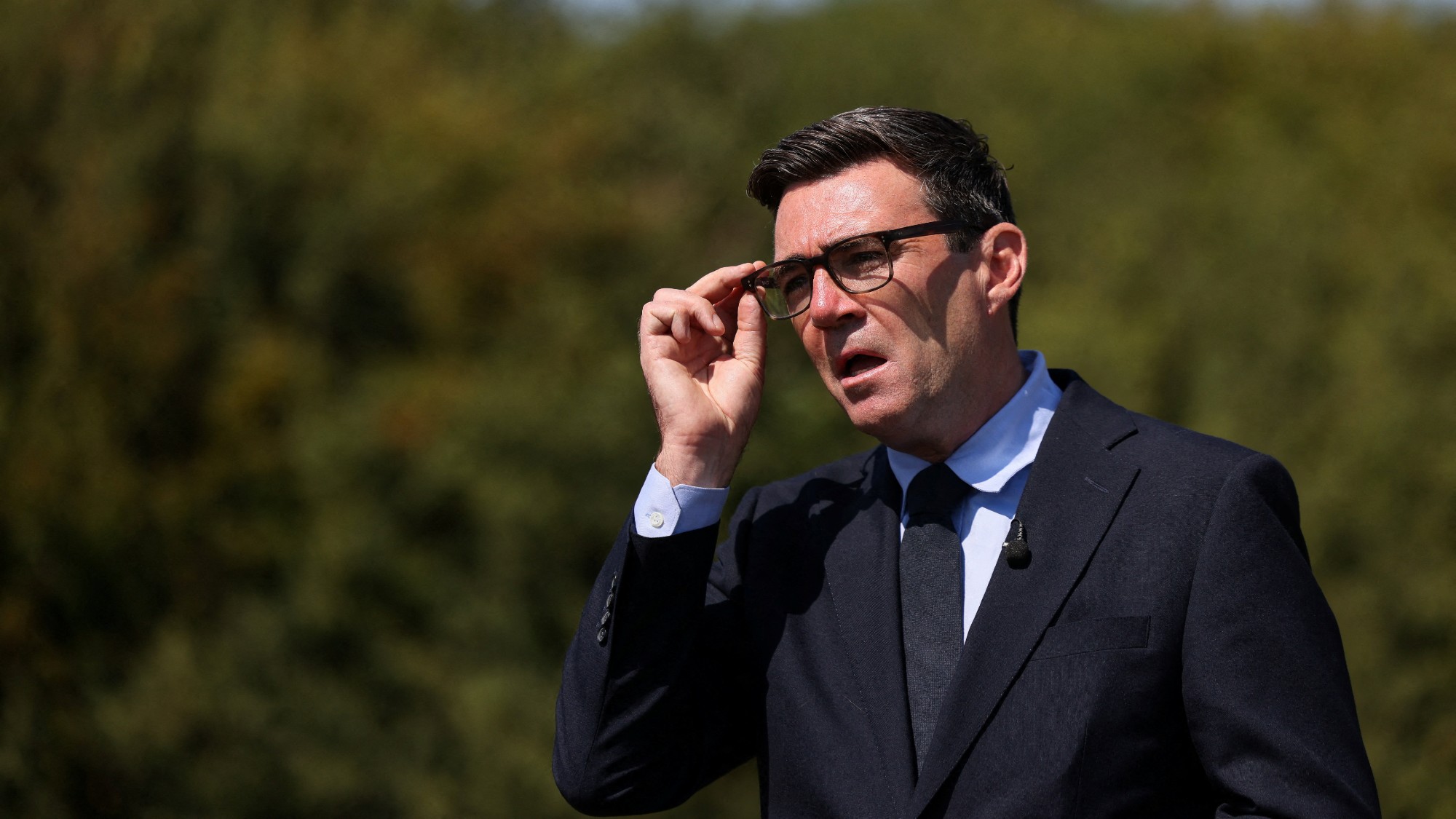 Andy Burnham adjusts his glasses while standing outdoors, against a backdrop of greenery