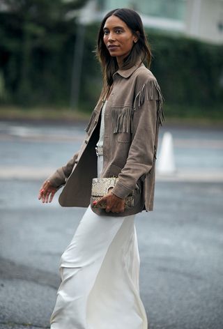 woman wearing tan fringe coat over white tank with white slip skirt