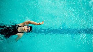 Woman doing backstroke in the swimming pool wearing black swimsuit and goggles