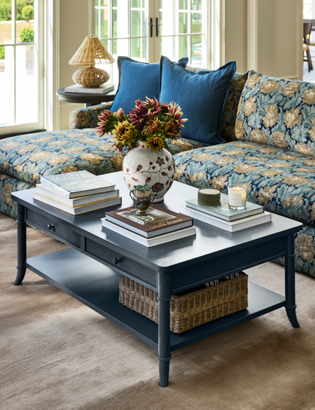 Two-tier navy blue coffee table with storage styled with stacks of coffee table books, a floral-patterned vase, match strike, and lit candles.