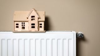 Modern radiator with wooden house near beige wall indoors. Winter heating efficiency