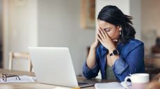 A woman puts her face in her hands while sitting in front of her laptop at a table.