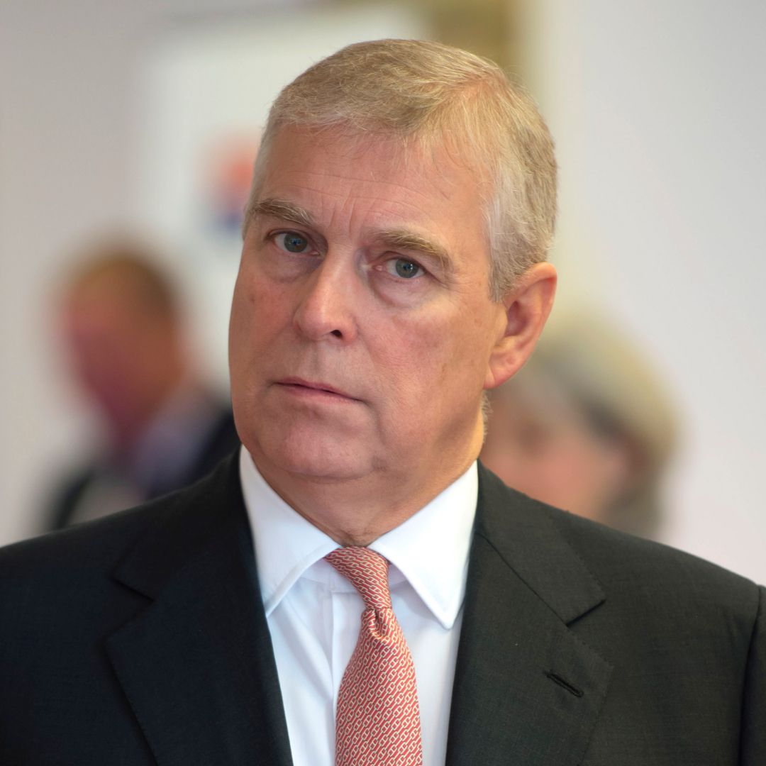 The former Prince Andrew looks annoyed while wearing a black suit, a white shirt, and a red tie