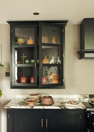 close up of a glass fronted kitchen cabinet, the wood work is painted a dark bluey black, matching the cabinets beneath. The worktop is a white marble with black veining