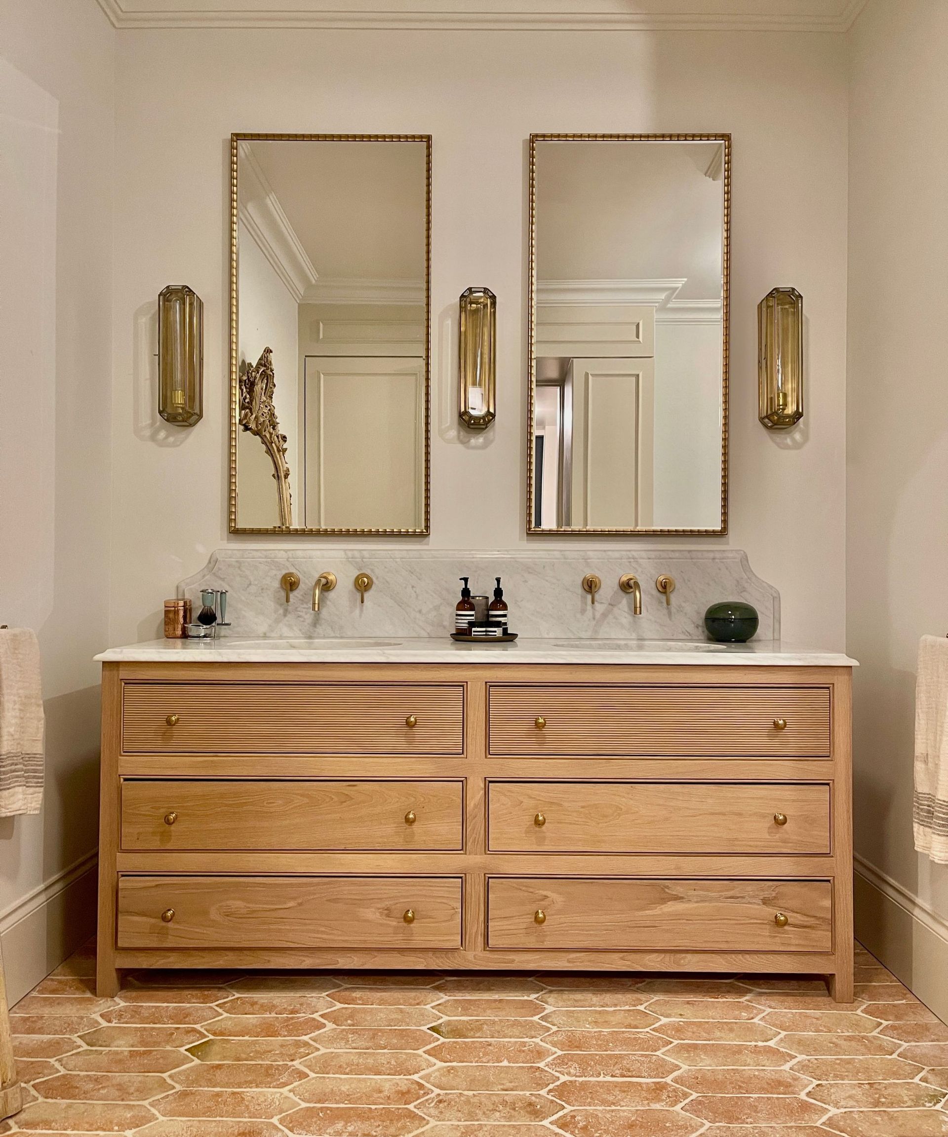 A classic bathroom with a wood vanity, gold mirrors, gold lighting, and terracotta floor tiles
