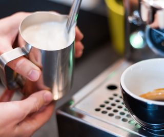 A jug of milk being textured using a steam wand with a cup of coffee on the right hand side
