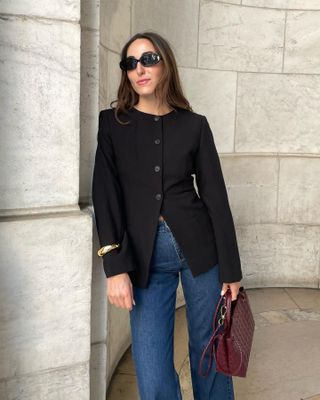 Fashion editor Anna LaPlaca poses in black oversize oval sunglasses, a black Aligne button-down crewneck blazer, a gold cuff, high-waisted jeans, and burgundy woven Bottega Veneta bag.