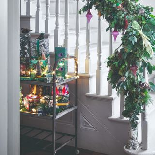 A hallway with a white staircase decorated with a garland and a trolley with gifts adorned with Christmas lights