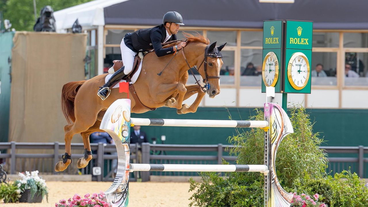 Showjumper Victor Bettendorf wears a black jacket and hat with white breeches on a chestnut horse called Qwando Van De Rispen, clears a large jump of black and white poles at Royal Windsor Horse Show