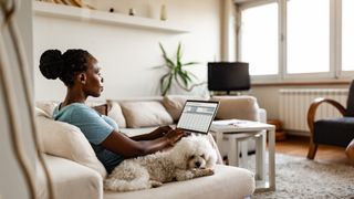 a woman uses a labtop while a small senior poodle mix lies next to her on a couch