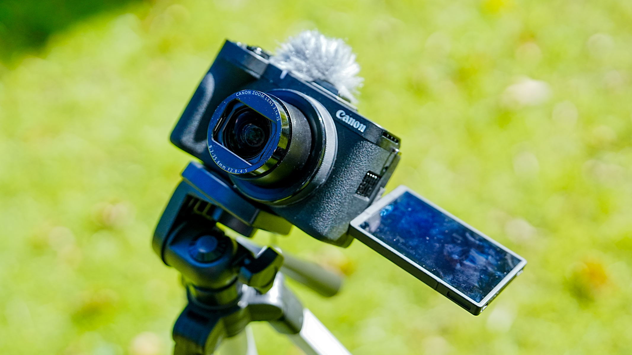 A Canon Powershot V1 with shutter open on a tripod with grass in the background.