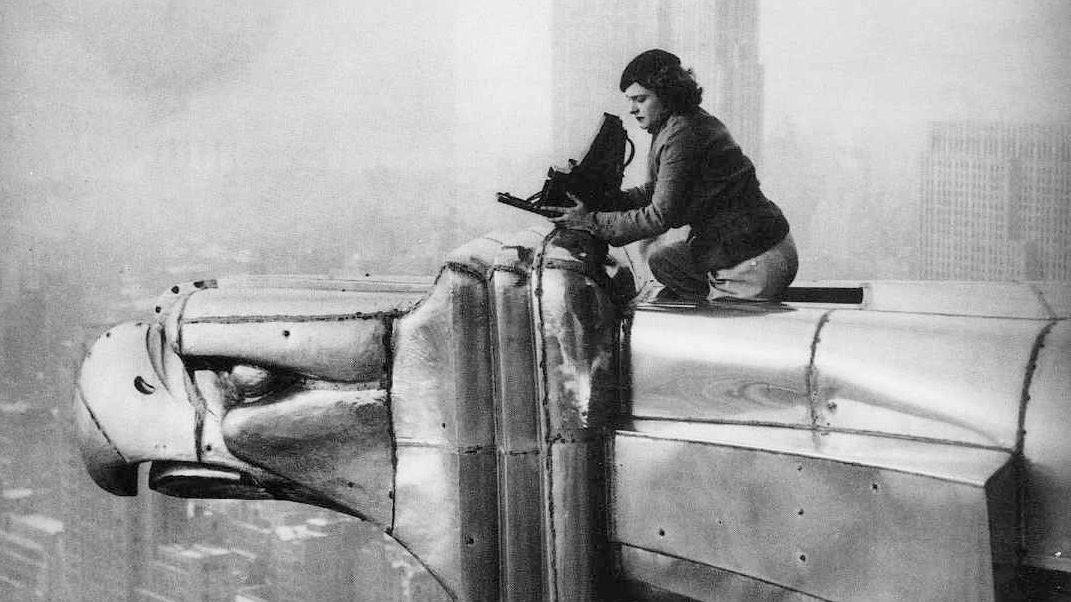 A woman in the 1930s atop a skyscraper, perched on a large eagle-shaped sculpture, operates a camera