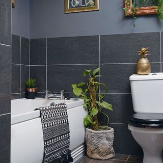 Black bathroom with white bath, black toilet seat, monochrome bath mat, black tiles, house plant