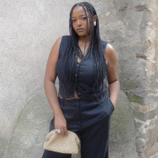 Woman in black v-neck vest, black trouser, raffia clutch bag, and silver earring against stone wall