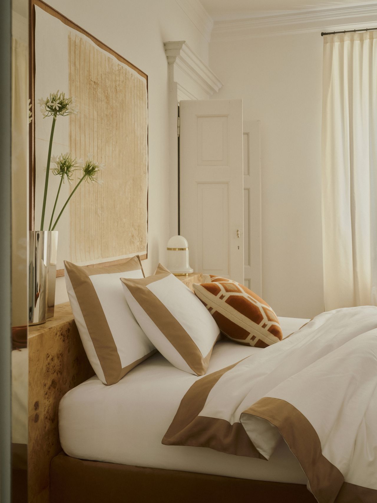 Image of an off-white bedroom with a luxury bed that has white bedding with tan trim. There is a red and white geometric throw pillow.
