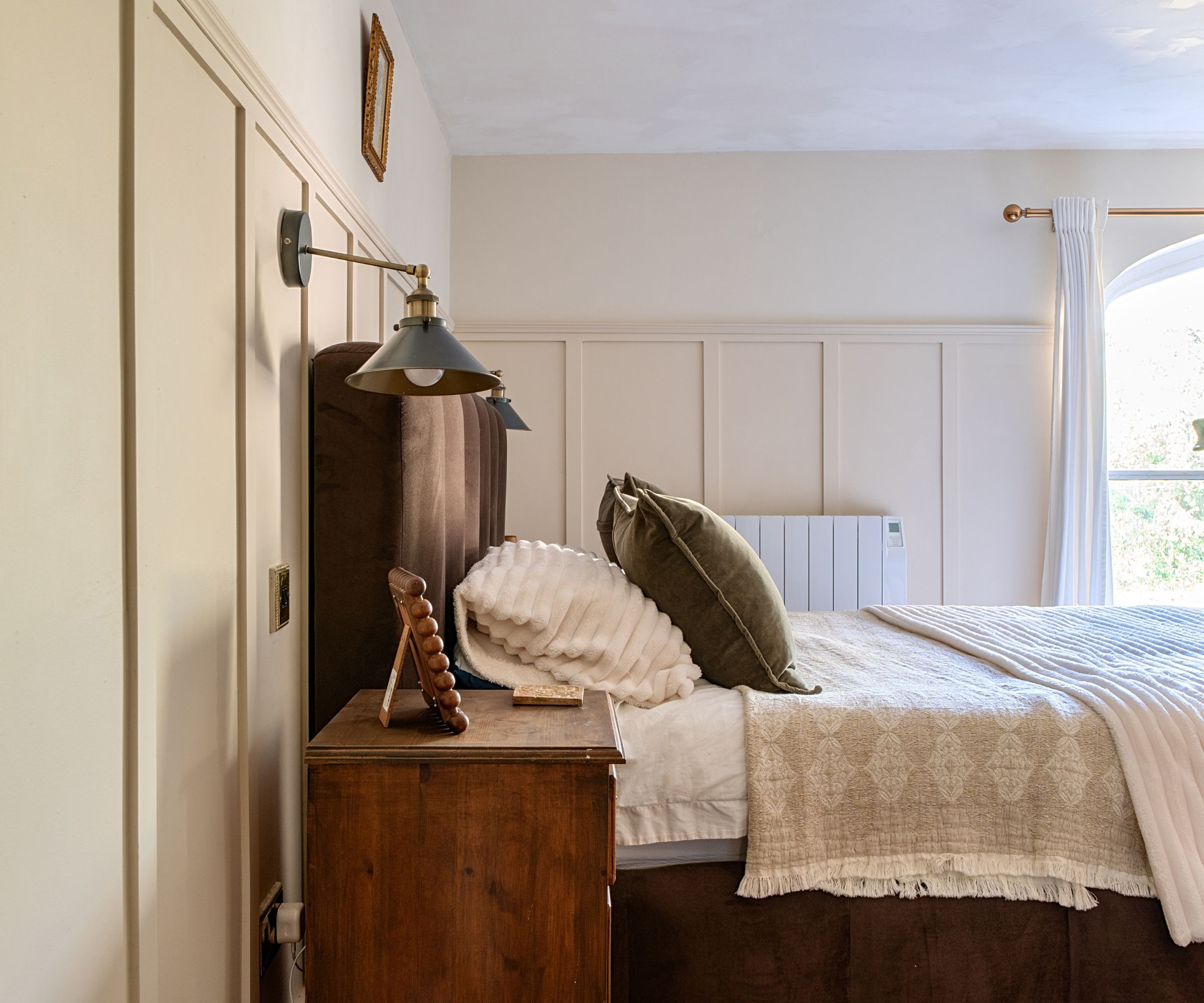 Cream panelled bedroom wall behind a bed with layered neutral bedding, olive cushion, wooden nightstand and brass wall lamp.