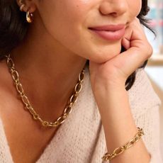 A happy woman wearing a thick, gold chain necklace and bracelet, as well as thick gold hoops from Peoples Jewellers.