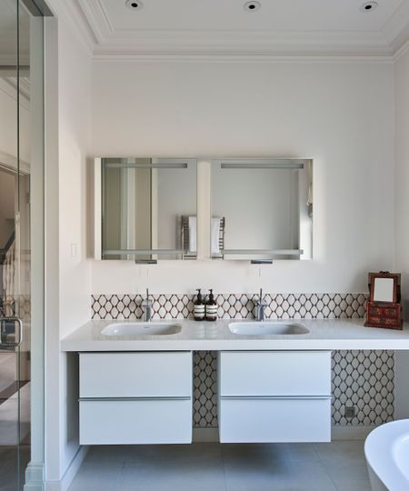 A bright bathroom with white basins with tiled backsplash, mirrored cabinets and a glass shower screen