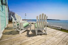 Three white chairs sit empty on a house's deck overlooking the ocean or beach.