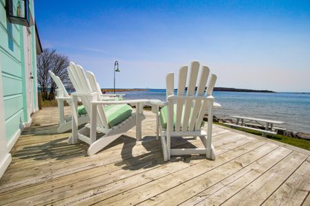 Three white chairs sit empty on a house's deck overlooking the ocean or beach.