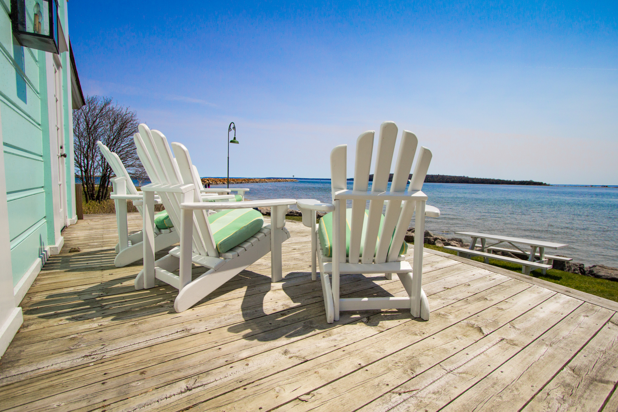 Three white chairs sit empty on a house's deck overlooking the ocean or beach.