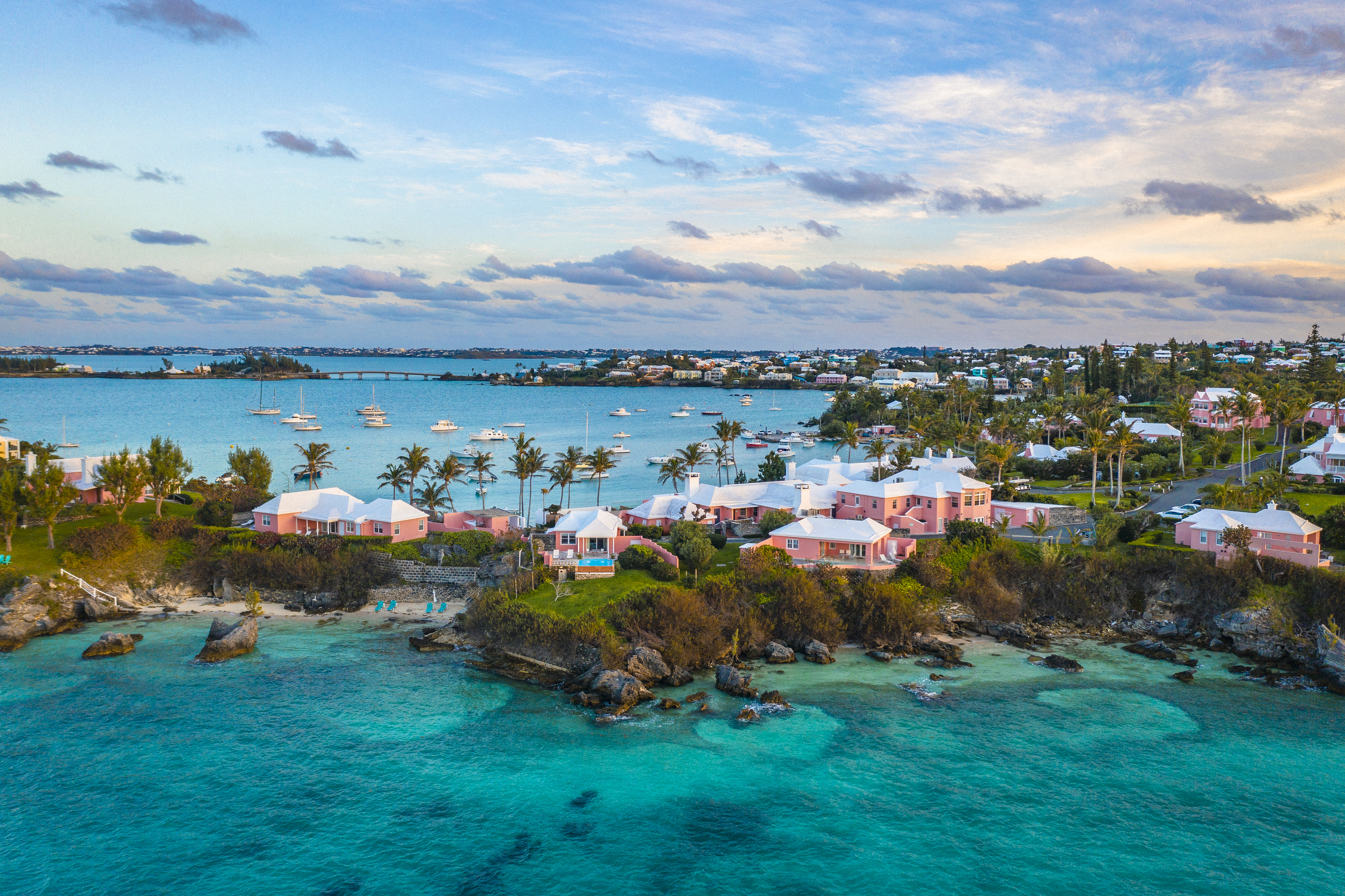 Tropical island coastline with pink houses and bright blue sea and sky. Some small beaches in the foreground and some boats and a bridge in the background.