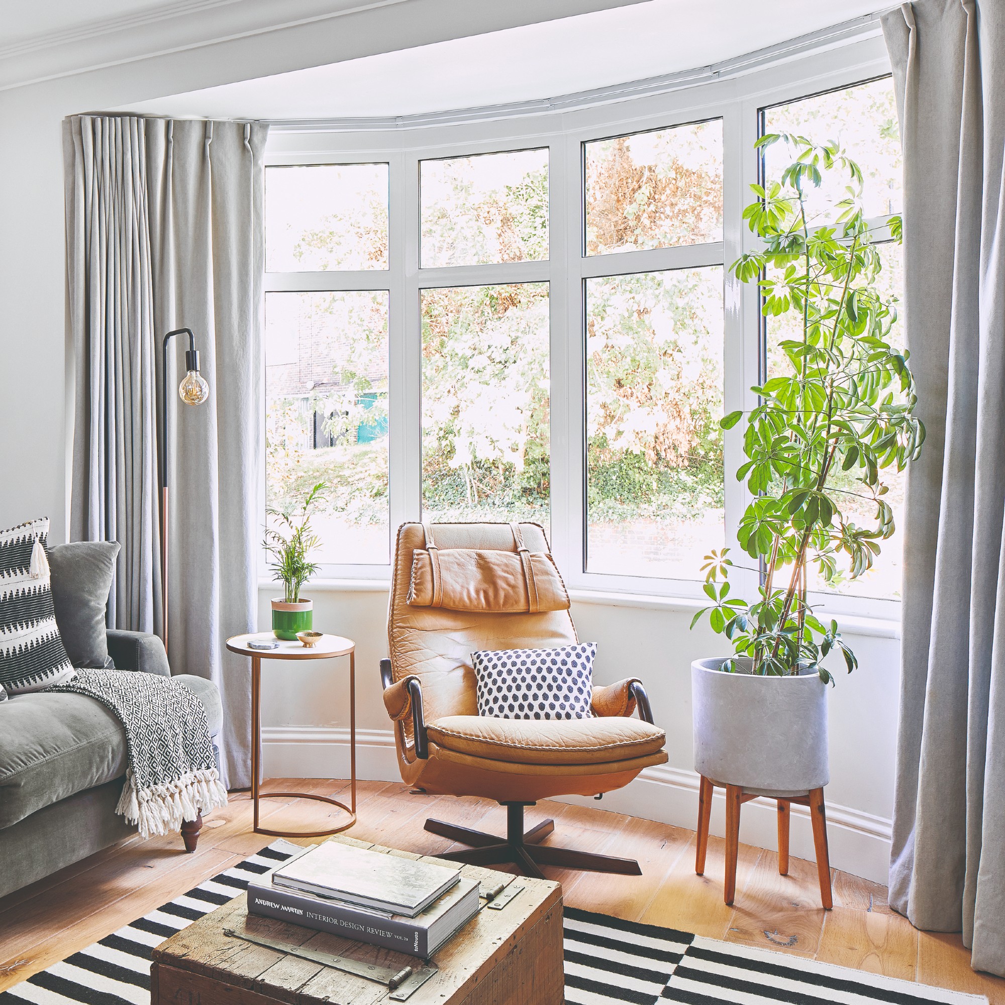 A living room with a bay window dressed with floor-length grey pleated curtains and adorned with houseplants and a swivel chair