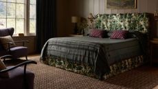 Moody, richly layered bedroom featuring a neutral heritage patterned rug, tapestry-print bed frame flanked by two ornately gilded candle sconces, and eclectic bedding.