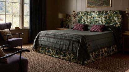 Moody, richly layered bedroom featuring a neutral heritage patterned rug, tapestry-print bed frame flanked by two ornately gilded candle sconces, and eclectic bedding.